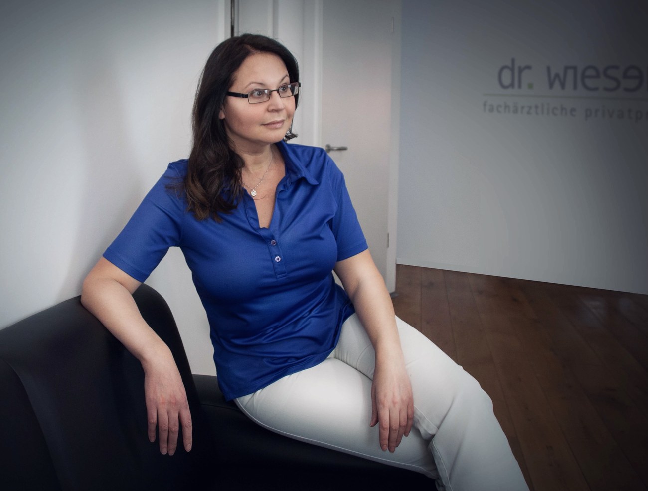 Dr. med. Natalie Wiesen im blauen Oberteil in ihrer Praxis, im Hintergrund das Praxislogo